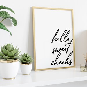 May include: A gold framed print with the text "hello sweet cheeks" in black cursive script. The print is on a white shelf with two potted succulents and a diffuser.