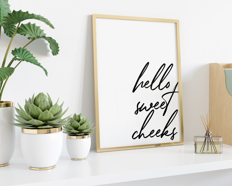 May include: A gold framed print with the text "hello sweet cheeks" in black cursive script. The print is on a white shelf with two potted succulents and a diffuser.