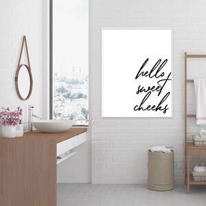 May include: A white bathroom with a framed print that says "hello sweet cheeks" hanging on the wall. There is a wooden ladder with towels hanging on it, a white sink with a mirror above it, and a brown wooden countertop.