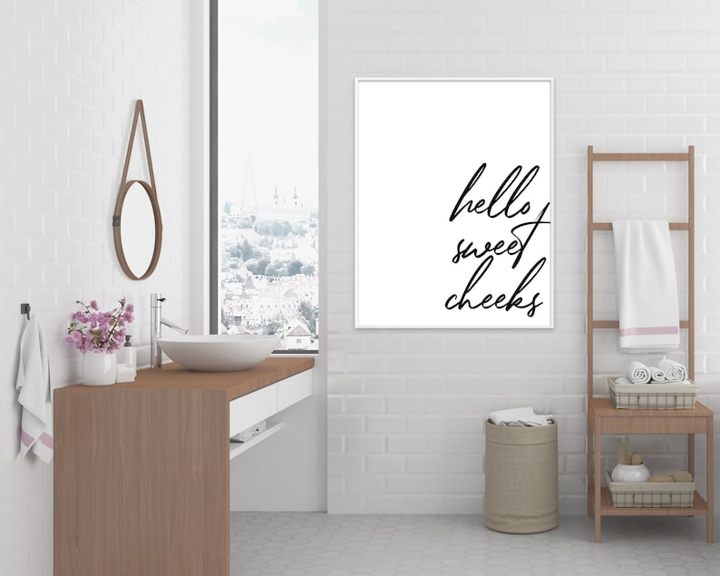 May include: A white bathroom with a framed print that says "hello sweet cheeks" hanging on the wall. There is a wooden ladder with towels hanging on it, a white sink with a mirror above it, and a brown wooden countertop.