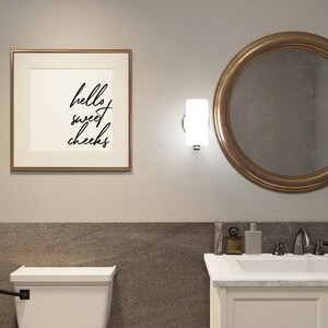 May include: A white bathroom with a toilet, a sink, and a mirror. The mirror has a gold frame. There is a framed print on the wall that says "hello sweet cheeks".
