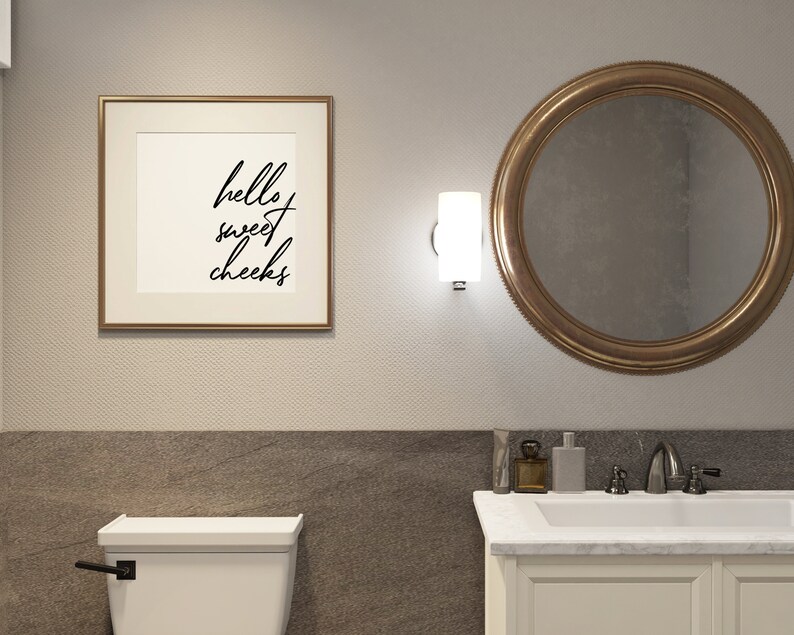 May include: A white bathroom with a toilet, a sink, and a mirror. The mirror has a gold frame. There is a framed print on the wall that says "hello sweet cheeks".