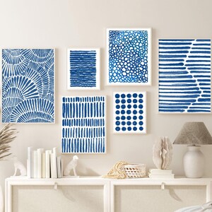 May include: A white dresser with six drawers, two on each side and two in the middle, with a white ceramic lamp on the right side. The dresser is decorated with five blue and white abstract art prints, and a white wicker basket sits in front of the dresser. A white bed with a white duvet cover is in the background.