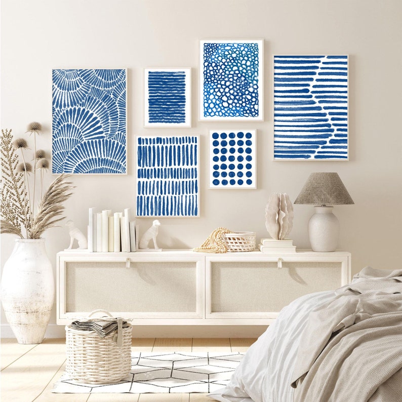 May include: A white dresser with six drawers, two on each side and two in the middle, with a white ceramic lamp on the right side. The dresser is decorated with five blue and white abstract art prints, and a white wicker basket sits in front of the dresser. A white bed with a white duvet cover is in the background.