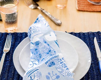May include: A table setting featuring a folded blue and white floral napkin on a white plate. A blue woven placemat, silverware, and a glass of water are also visible. The setting suggests a casual dining experience.