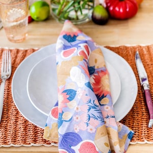 May include: A folded floral napkin in shades of lavender, pink, and orange rests on a white plate. A fork and knife are on either side of the plate, set on a woven placemat. The napkin features a colorful floral pattern.