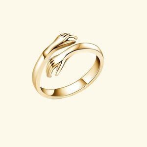 May include: A gold ring featuring two hands clasped together, forming a circle. The ring is open and adjustable.