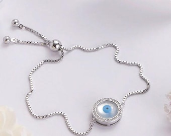 May include: A silver chain bracelet with an adjustable sliding clasp. The charm is a round pendant with a blue evil eye design surrounded by clear rhinestones.