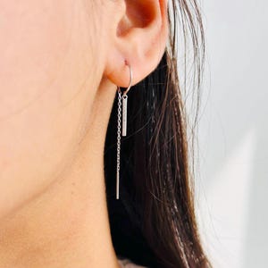 Puede incluir: Un pendiente plateado con un pequeño aro que se sujeta a la oreja. Una pequeña barra rectangular con cristales incrustados cuelga del aro, conectada a una barra más larga y colgante. El pendiente está diseñado para ser usado en una oreja.
