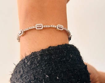 Op de afbeelding: Een zilveren armband met rechthoekige, heldere edelstenen. De armband is versierd met talrijke kleine, ronde edelstenen en heeft drie grotere rechthoekige stenen. De armband wordt om de pols gedragen, met een zwarte, getextureerde mouw op de achtergrond.