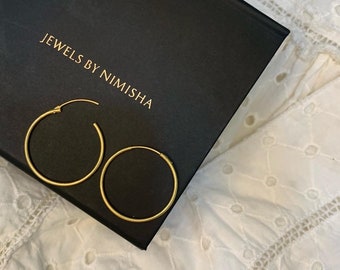 May include: Two gold hoop earrings on a black box with the text "Jewels by Nimisha".