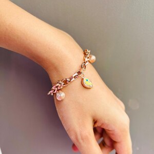 Puede incluir: Un brazalete de oro con un dije de corazón y otros dijes decorativos. El brazalete está en la muñeca de una persona.