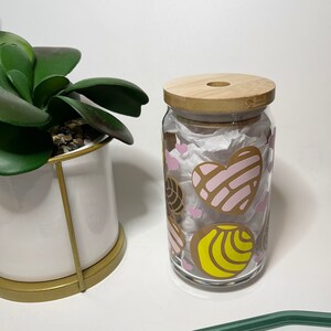May include: A glass drinking jar with a wooden lid and a green glass straw. The jar is decorated with pink, yellow, and brown hearts and swirls.