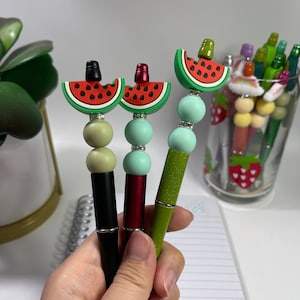 May include: Three pens with watermelon-shaped tops and colorful beads. The pens are black, red, and green.