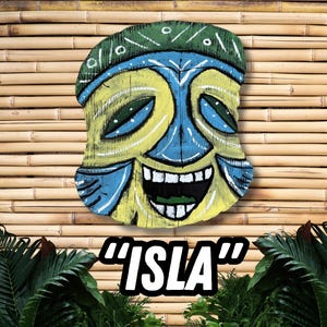 May include: A hand-painted wooden tiki mask with a smiling face, featuring yellow, blue, and green colors. The mask is mounted on a bamboo background, with the word "ISLA" in a black and white font at the bottom.