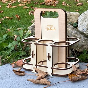 Puede incluir: Portabotellas de cerveza de madera para seis botellas con asa. El portabotellas es de madera clara y tiene una mancha marrón. La parte delantera del portabotellas tiene el texto "Herren Handtasche" en letras doradas.