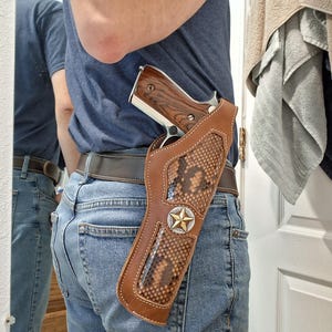 May include: A brown leather holster with a snakeskin pattern and a silver and gold star emblem. The holster is designed to be worn on a belt and holds a handgun with a wooden grip.
