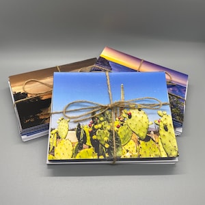 May include: A stack of landscape-themed greeting cards tied with twine. The cards feature images of cacti, sunsets, and other scenic views. The cards are square and have a white border. The twine is tied in a bow.
