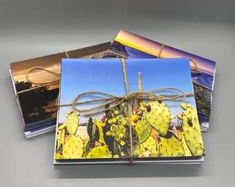 May include: A collection of landscape greeting cards bound with twine. The cards display images of cacti, sunsets, and other scenic views. The cards are square and have a white border. The twine is tied in a bow.