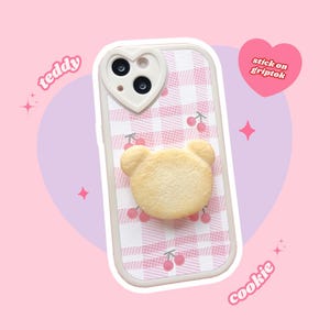 May include: A pink and white checked phone case with a heart-shaped camera cutout and a 3D cookie bear griptok. The griptok is shaped like a bear and has a light brown colour. The case has a pink and white checked pattern with small pink cherries printed on it. The text "stick on griptok" is printed in a heart shape on the right side of the image. The text "teddy" is printed on the left side of the image and the text "cookie" is printed on the right side of the image.