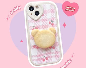 May include: A pink and white checkered phone case with a heart-shaped camera cutout and a 3D cookie bear griptok. The griptok is shaped like a bear and has a light brown color. The case has a pink and white checkered pattern with small pink cherries printed on it. The text "stick on griptok" is printed in a heart shape on the right side of the image. The text "teddy" is printed on the left side of the image and the text "cookie" is printed on the right side of the image.