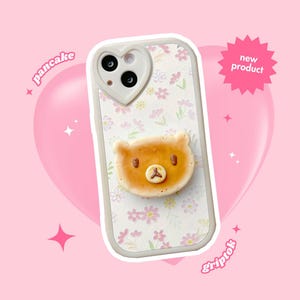 May include: A phone case with a light beige frame and a heart-shaped camera cutout. The case features a pancake-shaped bear design and a floral pattern. The words "pancake" and "griptok" are written on the image.