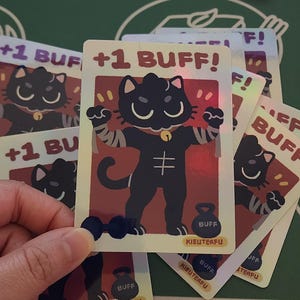 May include: A holographic trading card featuring a cartoon black cat flexing its muscles, with the text "+1 BUFF!". The card has a red and gold background, with a dumbbell and a weight labeled "BUFF". The card is part of a card game.