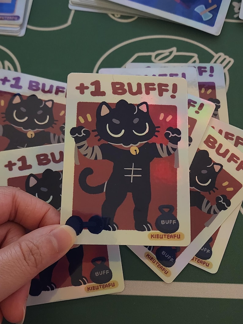 May include: A holographic trading card featuring a cartoon black cat flexing its muscles, with the text "+1 BUFF!". The card has a red and gold background, with a dumbbell and a weight labeled "BUFF". The card is part of a card game.