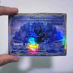 May include: A holographic trading card featuring the text "BARON PIT TOKEN BATTLEFIELD" with a cartoonish black creature. The card has a purple and blue background with mountains and text that reads "Units can move here from anywhere."