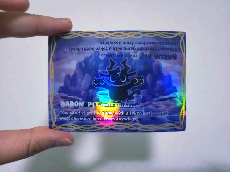 May include: A holographic trading card featuring the text "BARON PIT TOKEN BATTLEFIELD" with a cartoonish black creature. The card has a purple and blue background with mountains and text that reads "Units can move here from anywhere."