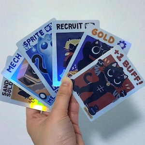 Puede incluir: Una mano sosteniendo un abanico de cartas coleccionables con ilustraciones de gatos. Las cartas presentan texto como "SPRITE", "RECRUIT" y "GOLD", junto con otros gráficos. Las cartas tienen un brillo holográfico, agregando un elemento visual dinámico.