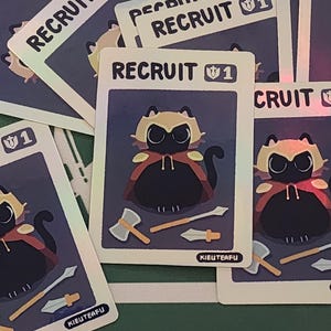 May include: A collection of trading cards featuring a cartoon cat character in a cape. The cards have the word "RECRUIT" at the top and include illustrations of weapons. The artist's name, "KIEUTEAFU", is printed on the bottom of the cards.