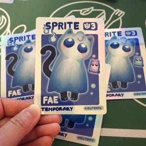 May include: Several trading cards featuring a blue cat-like creature with large blue eyes and a black tail. The cards have the text "SPRITE V3", "FAE TEMPORARY", and "KIEUTEAFU". The cards are on a green surface.