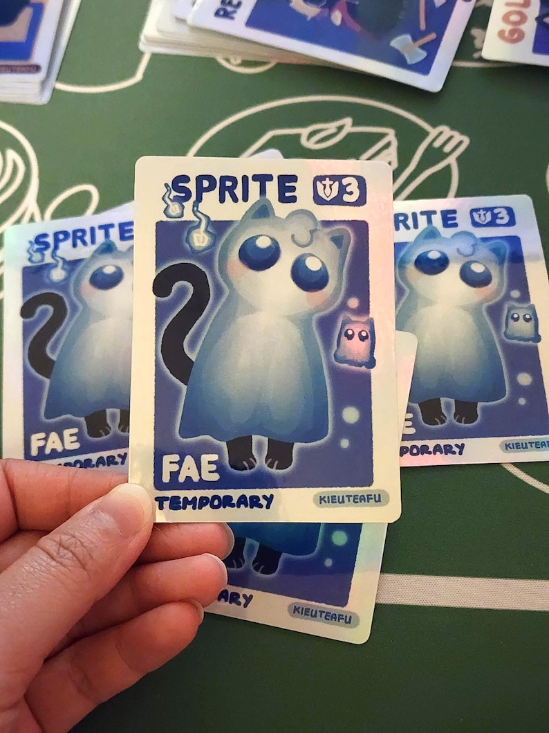 May include: Several trading cards featuring a blue cat-like creature with large blue eyes and a black tail. The cards have the text "SPRITE V3", "FAE TEMPORARY", and "KIEUTEAFU". The cards are on a green surface.