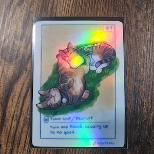 May include: A holographic trading card featuring two cartoon cats. One cat is lying on its back, and the other is resting on top. The card has text that reads "Token Unit/Recruit" and "Twix and Roche, usually up to no good."