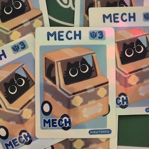 May include: Several trading cards featuring a cartoon cat driving an orange car. The cards have the word "MECH" in blue, and the cat has large, round eyes. The cards are on a green and white surface.