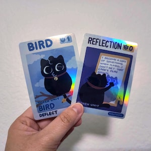 May include: Two trading cards are held in a hand. The card on the left says "BIRD" and features a black cat with large eyes. The card on the right says "REFLECTION" and has a black cat with a holographic effect. The cards have a blue background.