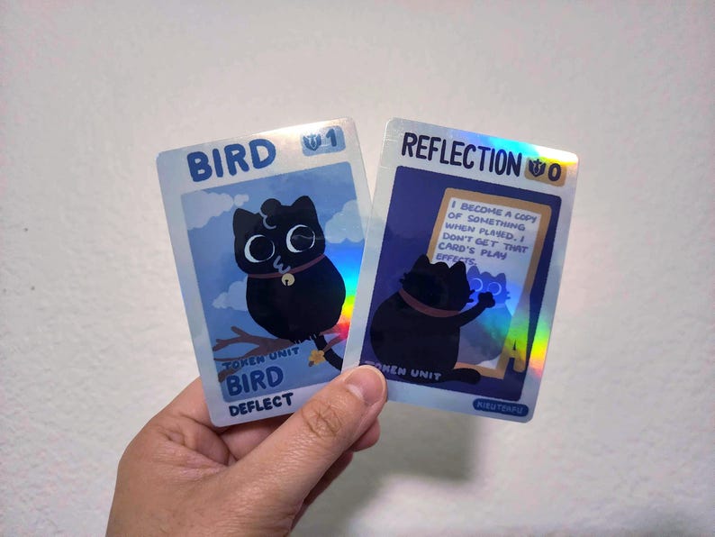 May include: Two trading cards are held in a hand. The card on the left says "BIRD" and features a black cat with large eyes. The card on the right says "REFLECTION" and has a black cat with a holographic effect. The cards have a blue background.