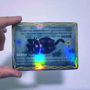 May include: A holographic trading card featuring a black cat illustration. The card has the word "BRUSH" and game instructions. The card is held by a hand and has a blue and gold border with floral accents.