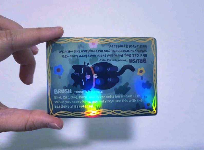 May include: A holographic trading card featuring a black cat illustration. The card has the word "BRUSH" and game instructions. The card is held by a hand and has a blue and gold border with floral accents.