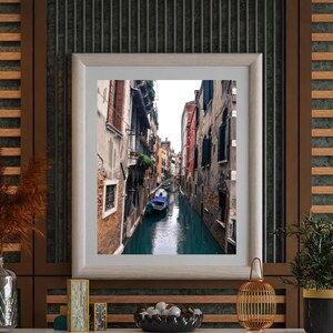 May include: A framed photograph of a narrow canal in Venice, Italy. The canal is lined with buildings on both sides, and a gondola is floating in the water. The photograph is displayed on a wooden shelf with other decorative items, including a vase with dried flowers, a small wooden box, and a bowl of decorative stones.