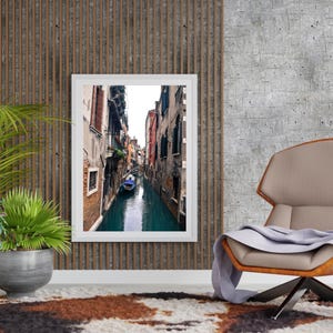 May include: A framed photograph of a narrow canal in Venice, Italy. The canal is lined with tall buildings and a small boat is visible in the water.