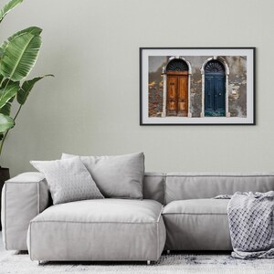 K&ouml;nnte beinhalten: Gerahmtes Foto mit zwei gew&ouml;lbten T&uuml;ren, eine braun und eine blaue, in einer verwitterten Wand. Das Bild hat einen Vintage-Look. Der Rahmen h&auml;ngt &uuml;ber einem grauen Sofa. Ein dekoratives Wandbild.
