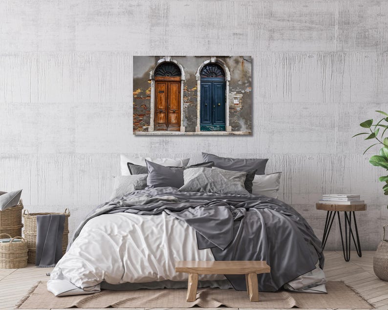 May include: A bedroom with a bed with white and gray bedding, a wooden bench, and a side table with books. A framed picture of two arched doorways with one door painted blue and the other painted brown is hanging on the wall behind the bed.