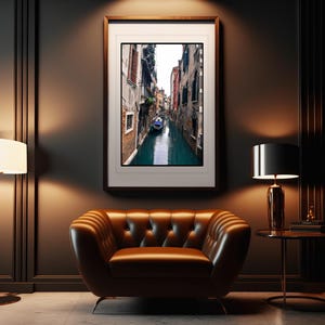 May include: A framed photograph of a narrow canal in Venice, Italy. The canal is lined with buildings on both sides, and a small boat is visible in the water. The photograph is in a dark wood frame and is hanging on a dark wall.