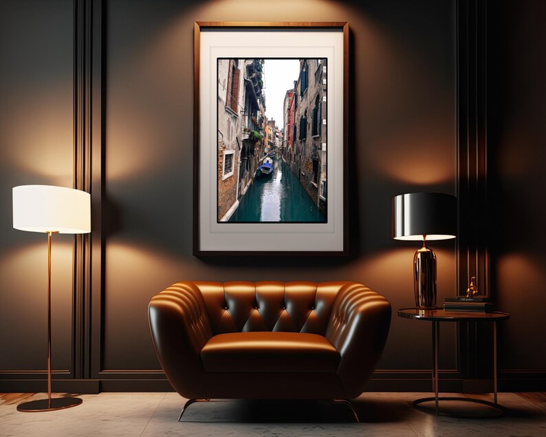 May include: A framed photograph of a narrow canal in Venice, Italy. The canal is lined with buildings on both sides, and a small boat is visible in the water. The photograph is in a dark wood frame and is hanging on a dark wall.