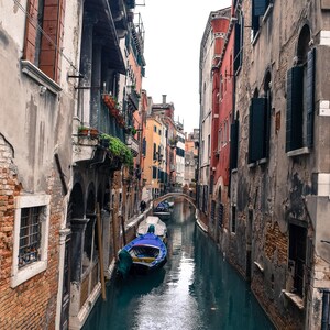 May include: A narrow canal in Venice, Italy, with old buildings on either side. A blue and white boat is docked in the canal.