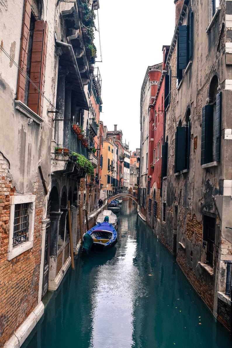May include: A narrow canal in Venice, Italy, with old buildings on either side. A blue and white boat is docked in the canal.