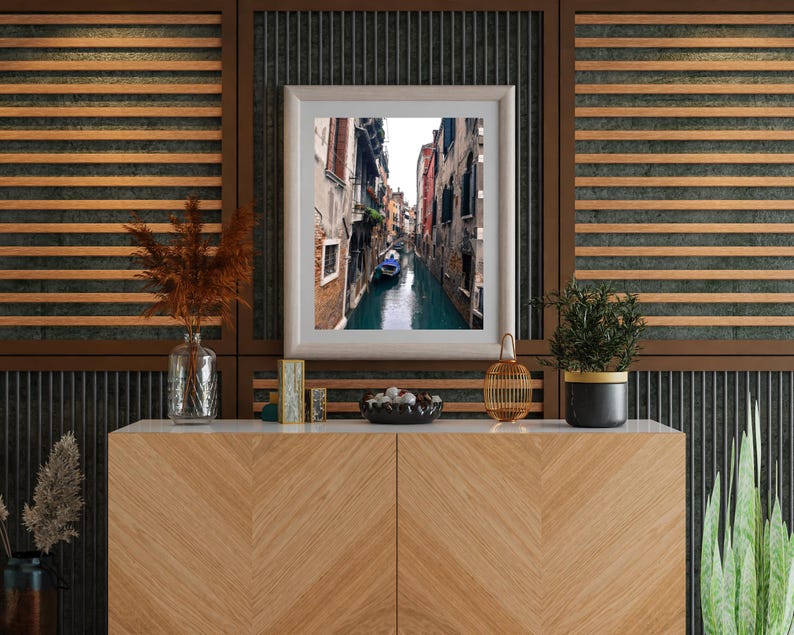 May include: A framed photograph of a narrow canal in Venice, Italy. The canal is lined with buildings on both sides, and a gondola is floating in the water. The photograph is displayed on a wooden shelf with other decorative items, including a vase with dried flowers, a small wooden box, and a bowl of decorative stones.