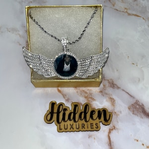 Può includere: Collana con pendente argentato con foto al centro, circondata da decorazioni a forma di ali con cristalli. La collana è esposta in una scatola dorata, con un cartello di legno che recita "Hidden LUXURIES".